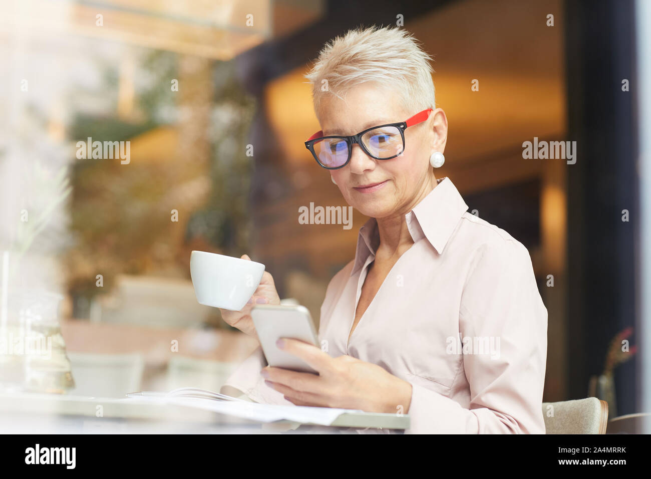 Femme mature avec de courts cheveux blonds en utilisant son téléphone portable tout en buvant du café pendant pause café dans cafe Banque D'Images