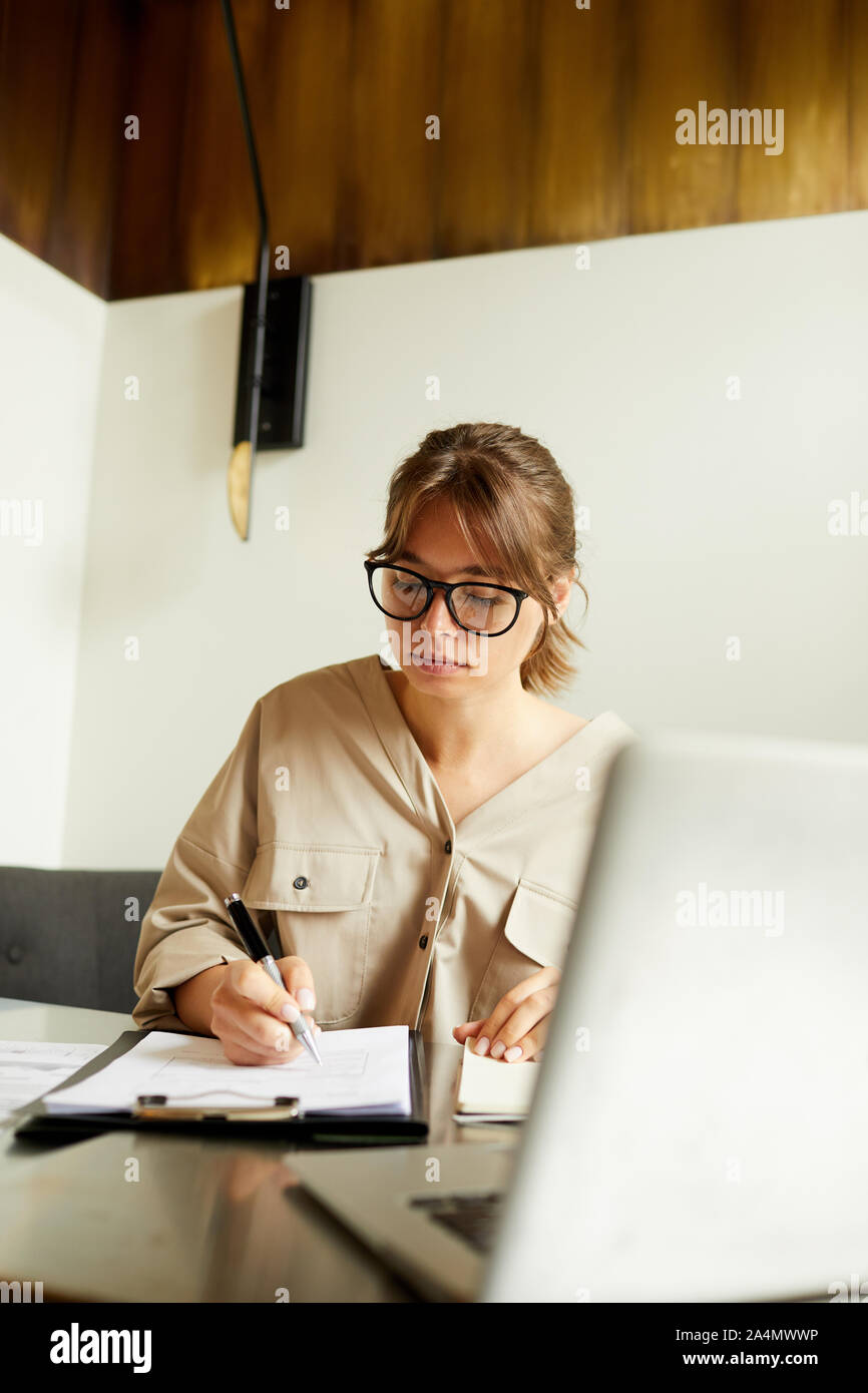 Concentrés young businesswoman wearing eyeglasses assis à la table en face de l'ordinateur portable et la signature de document Banque D'Images