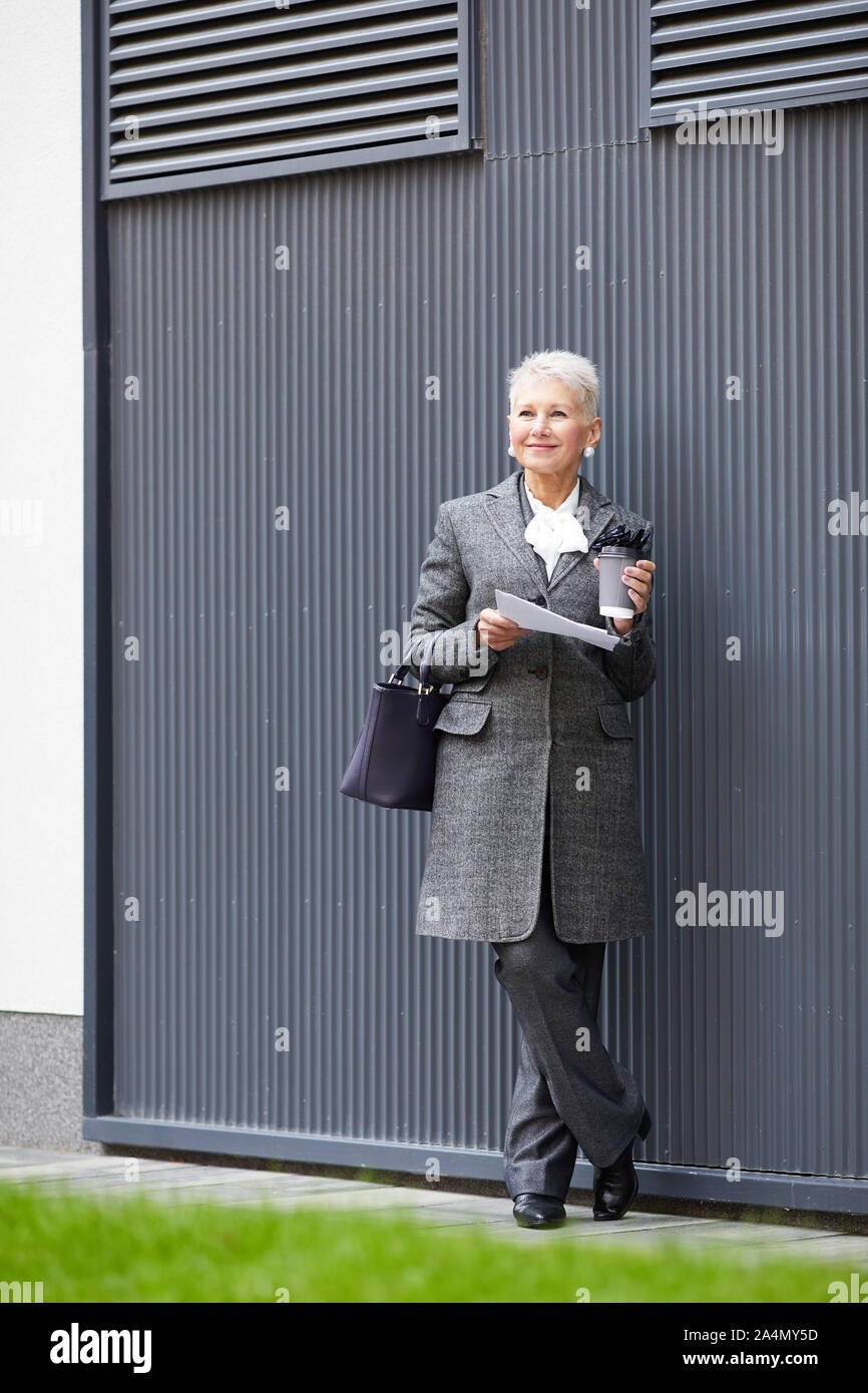 Femme mature blonde élégante avec des cheveux courts holding documents et boire du café dans ses mains tout en se tenant à l'extérieur Banque D'Images