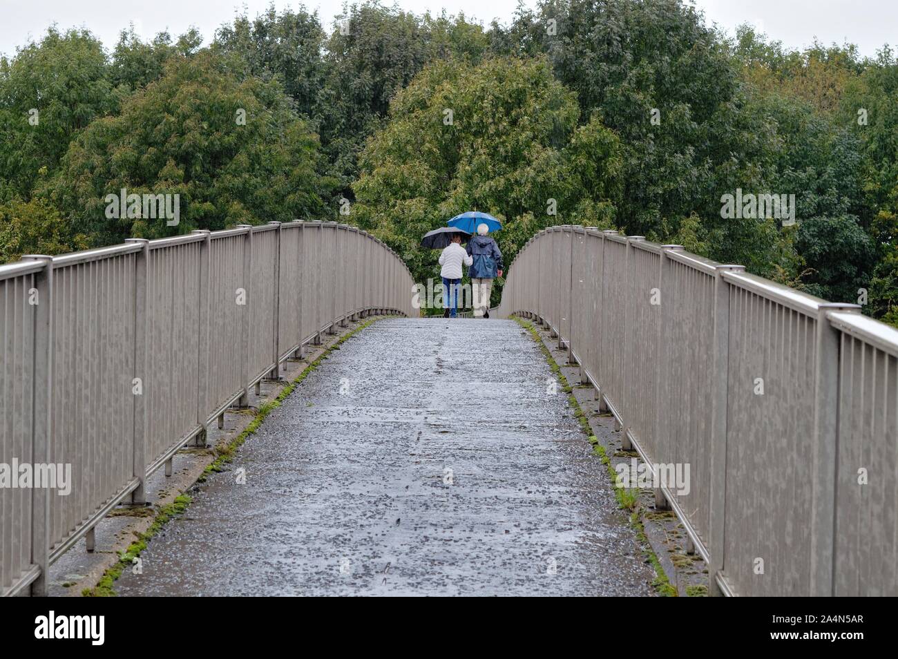Vue arrière d'un couple de personnes âgées avec parapluie marchant sur une passerelle dans la pluie, Shepperton Surrey England UK Banque D'Images