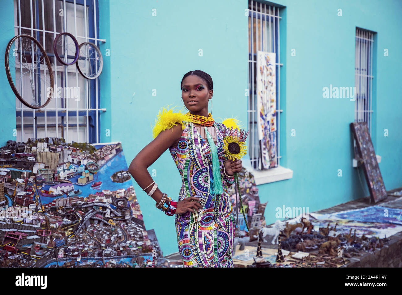 Une femme dans une robe longue aux portes d'une maison bleue en Bo Kaap, tenant un tournesol en perles Banque D'Images