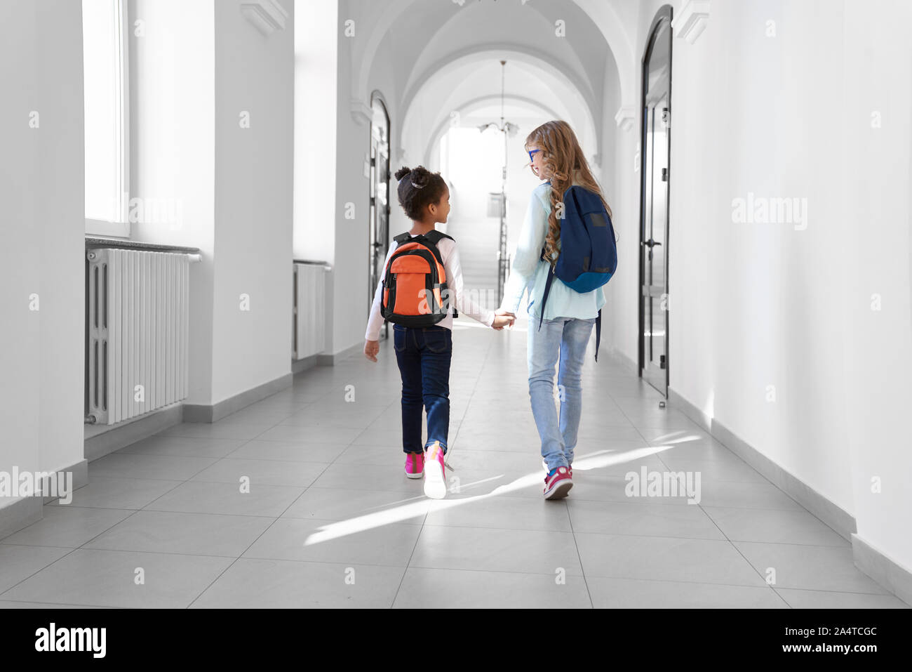 Deux écolières en jeans et baskets avec sacs à dos marchant dans la lumière à long couloir après les cours. Les amis se tenant la main et de parler à l'école. Banque D'Images