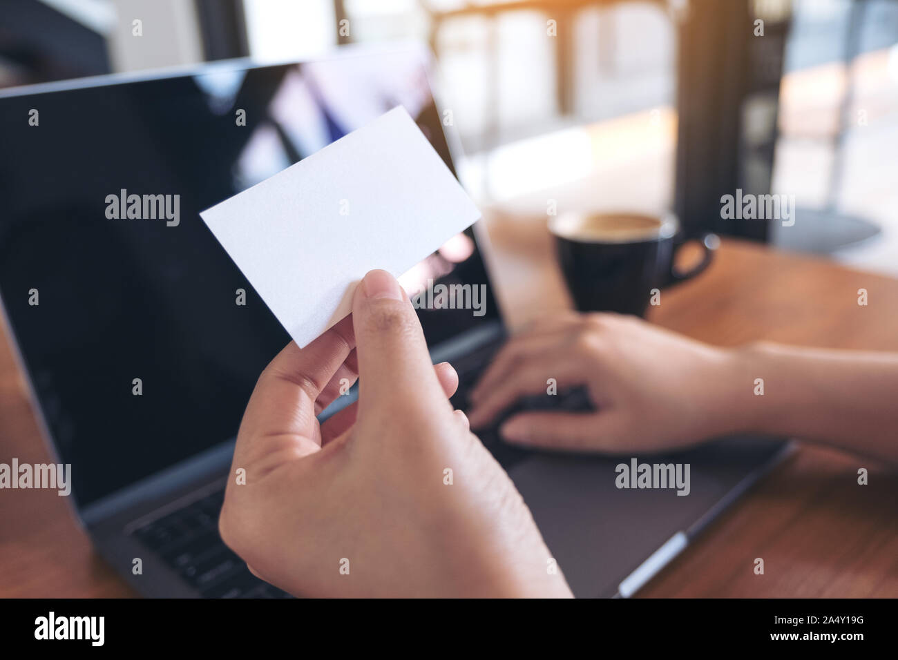 A hand holding vide blanc carte d'affaires tout en utilisant un ordinateur portable avec une tasse à café sur table en bois in office Banque D'Images