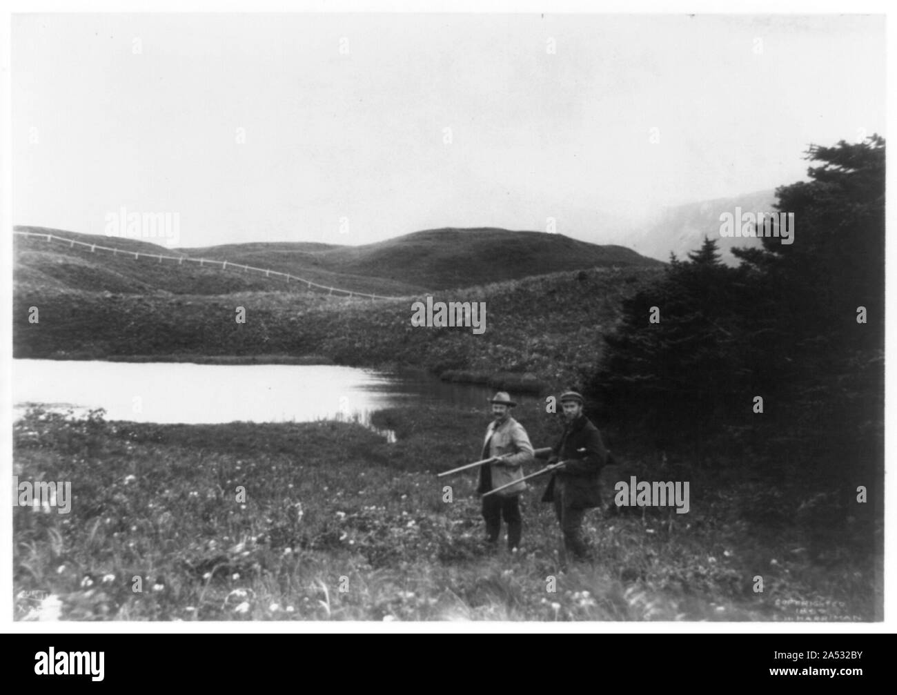 Deux hommes avec des fusils en premier plan, étang et montagne en arrière-plan, prises au cours de l'expédition Harriman en Alaska, 1899 Banque D'Images
