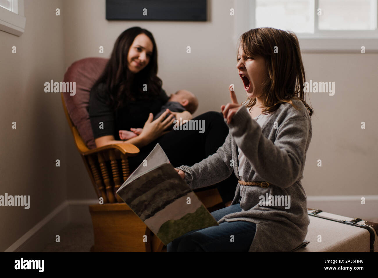 Girl with book pulling faces, alors que mère berceaux frère bébé dans la salle de séjour fauteuil Banque D'Images