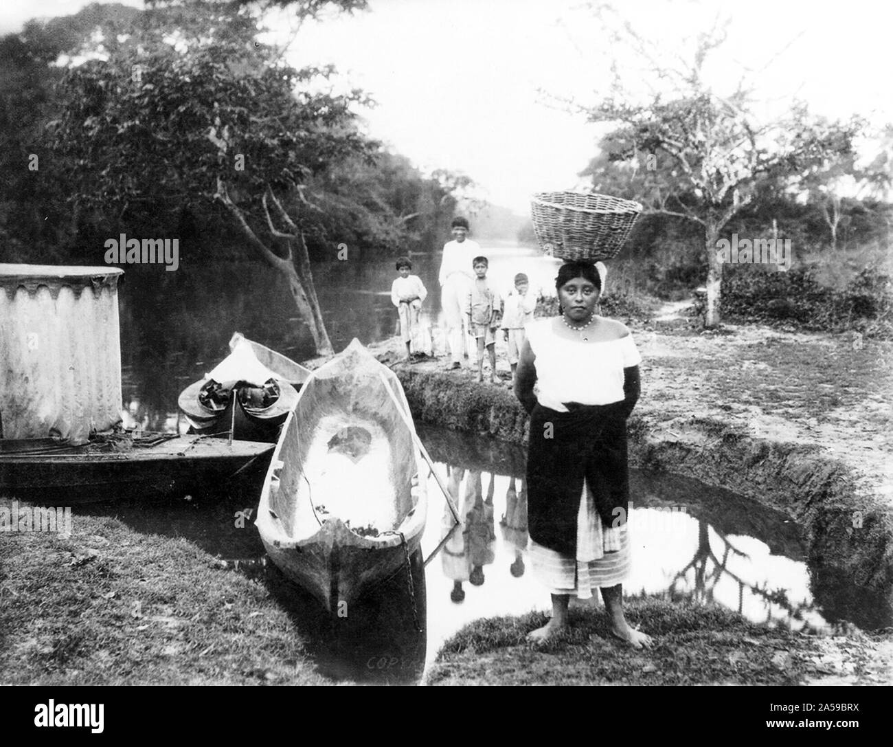 Femme avec panier sur la tête et 4 enfants debout sur banque ; 2 bateaux pirogue et 2 autres bateaux dans l'eau, 1917 Cuba Banque D'Images