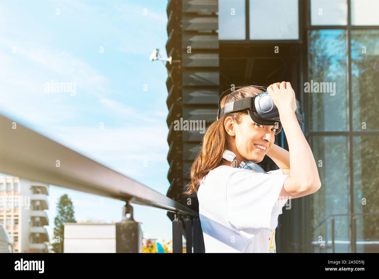 Belle jeune femme en t-shirt blanc mise sur casque de réalité virtuelle et à la recherche en appareil photo avec un grand sourire mignon. Entreprise moderne en verre buildi Banque D'Images