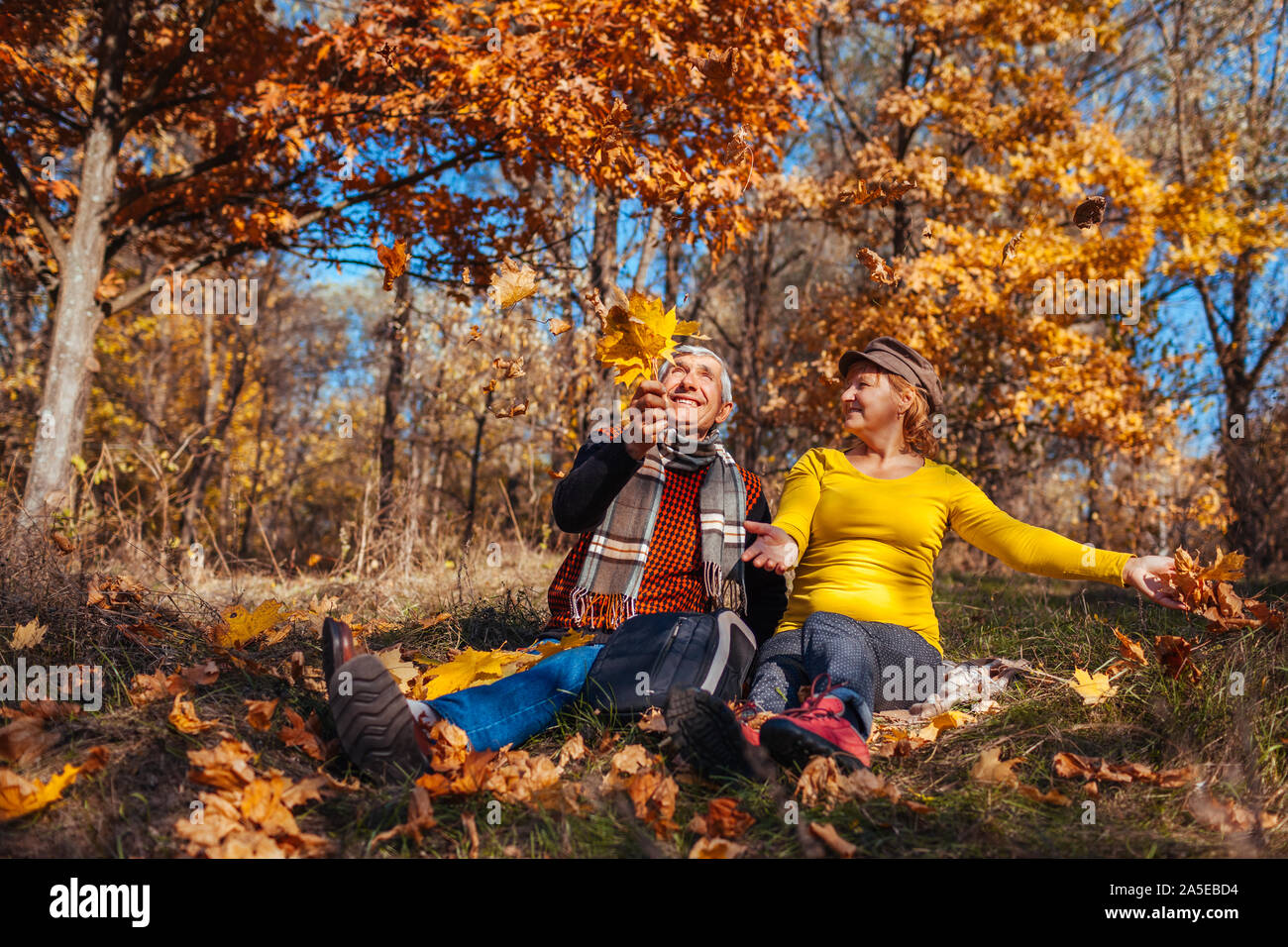 Saison d'automne de plaisir. Feuilles jeter Senior couple sitting in park. Heureux l'homme et de la femme de détente en plein air apprécier la nature Banque D'Images