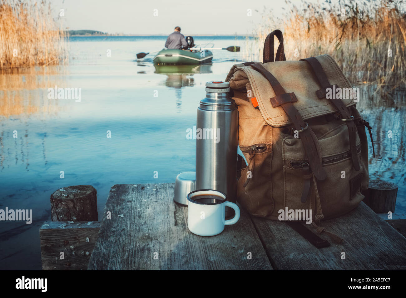 Mug émaillé de café ou de thé, sac à dos de voyageur et le thermos sur la jetée en bois sur le lac tranquille. Un pêcheur de bateau en caoutchouc en arrière-plan. Banque D'Images Mug émaillé de café ou de thé, sac à dos de voyageur et le thermos sur la jetée en bois sur le lac tranquille. Un pêcheur de bateau en caoutchouc en arrière-plan. Banque D'Images