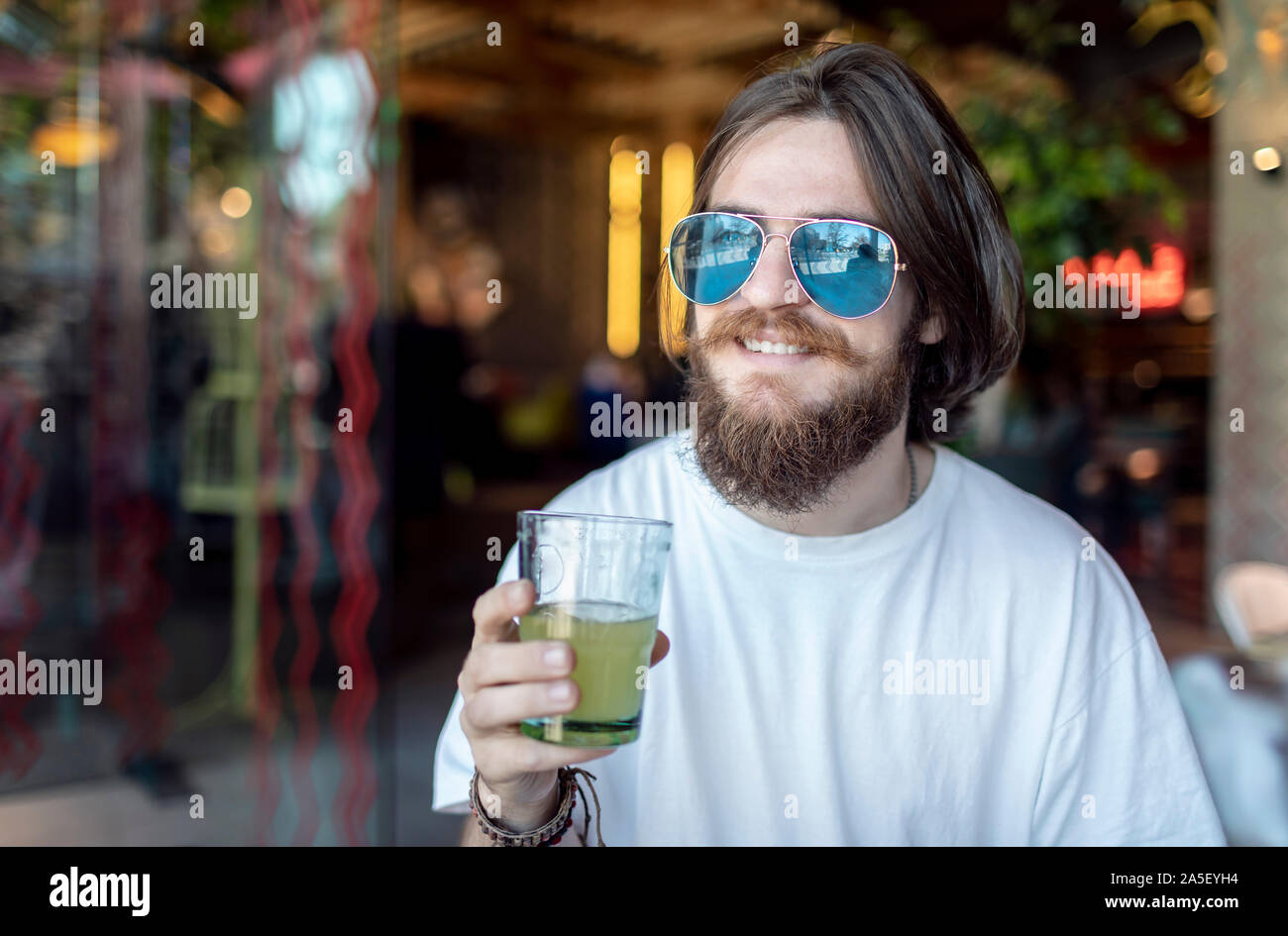Heureux jeune homme en blanc T-shirt et lunettes de soleil miroir apprécie son cocktail relance Banque D'Images