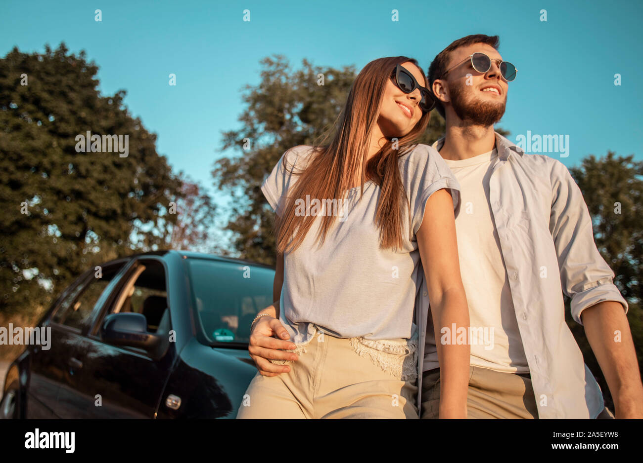 Couple aimant posant sur un coucher de soleil route de campagne appuyé contre un capot de voiture low angle view Banque D'Images