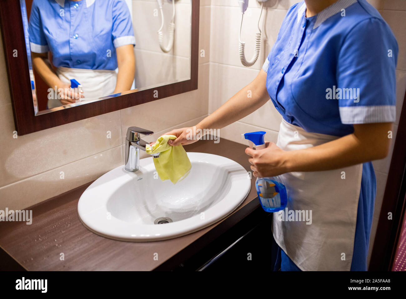 Jeune femme de chambre en uniforme, debout devant un miroir dans une salle de bains privative et un lavabo avec un détergent de nettoyage Banque D'Images