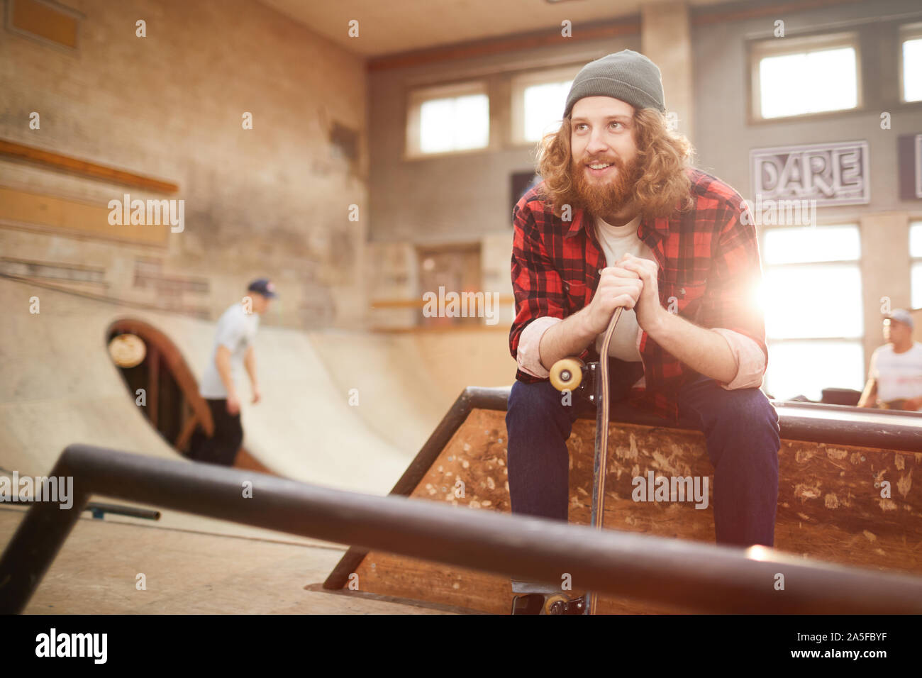 Portrait d'homme barbu contemporain à l'écart et souriant assis sur la rampe en skating park, copy space Banque D'Images