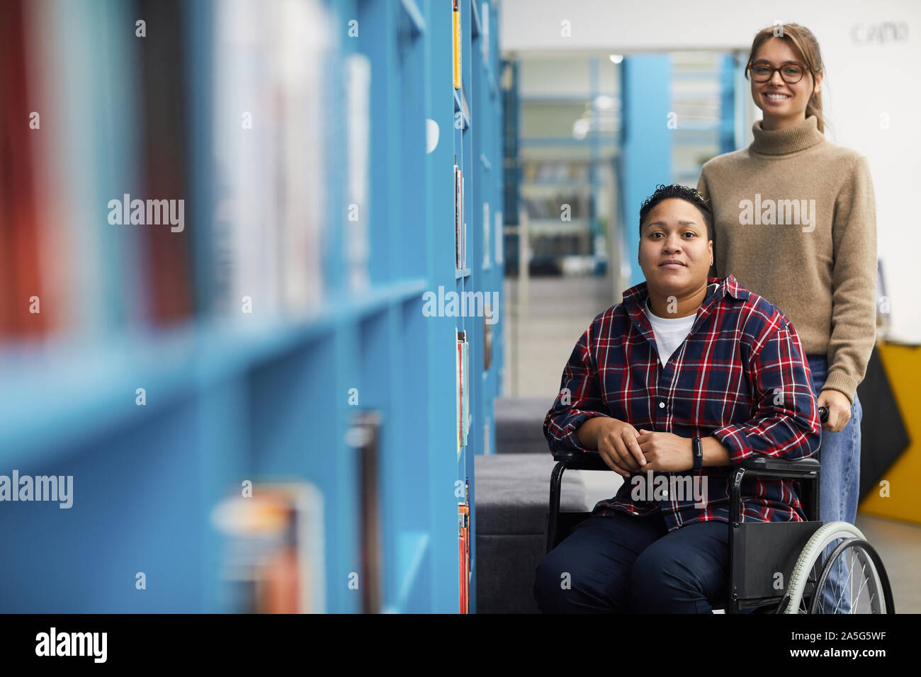 Portrait de deux étudiants de niveau collégial, l'un d'entre eux en fauteuil roulant, posant ainsi l'article par des rayons de bibliothèque, copy space Banque D'Images