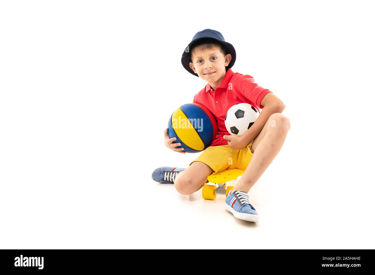 Un petit garçon au Panama, un maillot jaune, short rouge et blanc sneakers est assise sur un penny jaune et est titulaire d'un terrain de basket-ball, un ballon multicolore noir soccer Banque D'Images