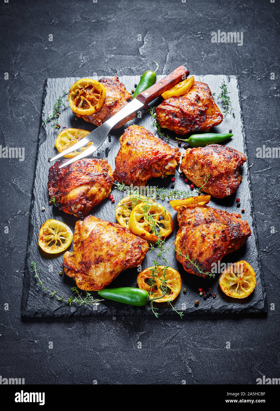 Close-up de cuisses de poulet frit croustillant avec des tranches de citron, thym et piments jalapeno sur une pierre noire sur un plateau de table en béton, vue d'abo Banque D'Images