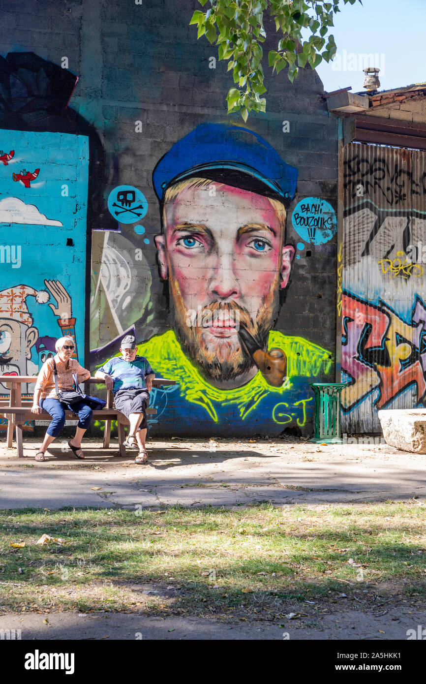 Un couple de retraités s'asseoir détendu sur un banc devant un mur art portrait - grafittee, Redon, France Banque D'Images