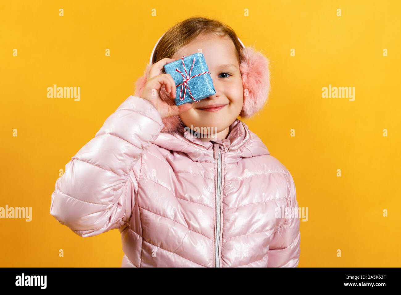 Petite fille joyeuse avec une petite boîte avec un cadeau. Un enfant dans une veste et casque chaud sur un fond jaune. Le concept de Noël, Nouvel An, Banque D'Images