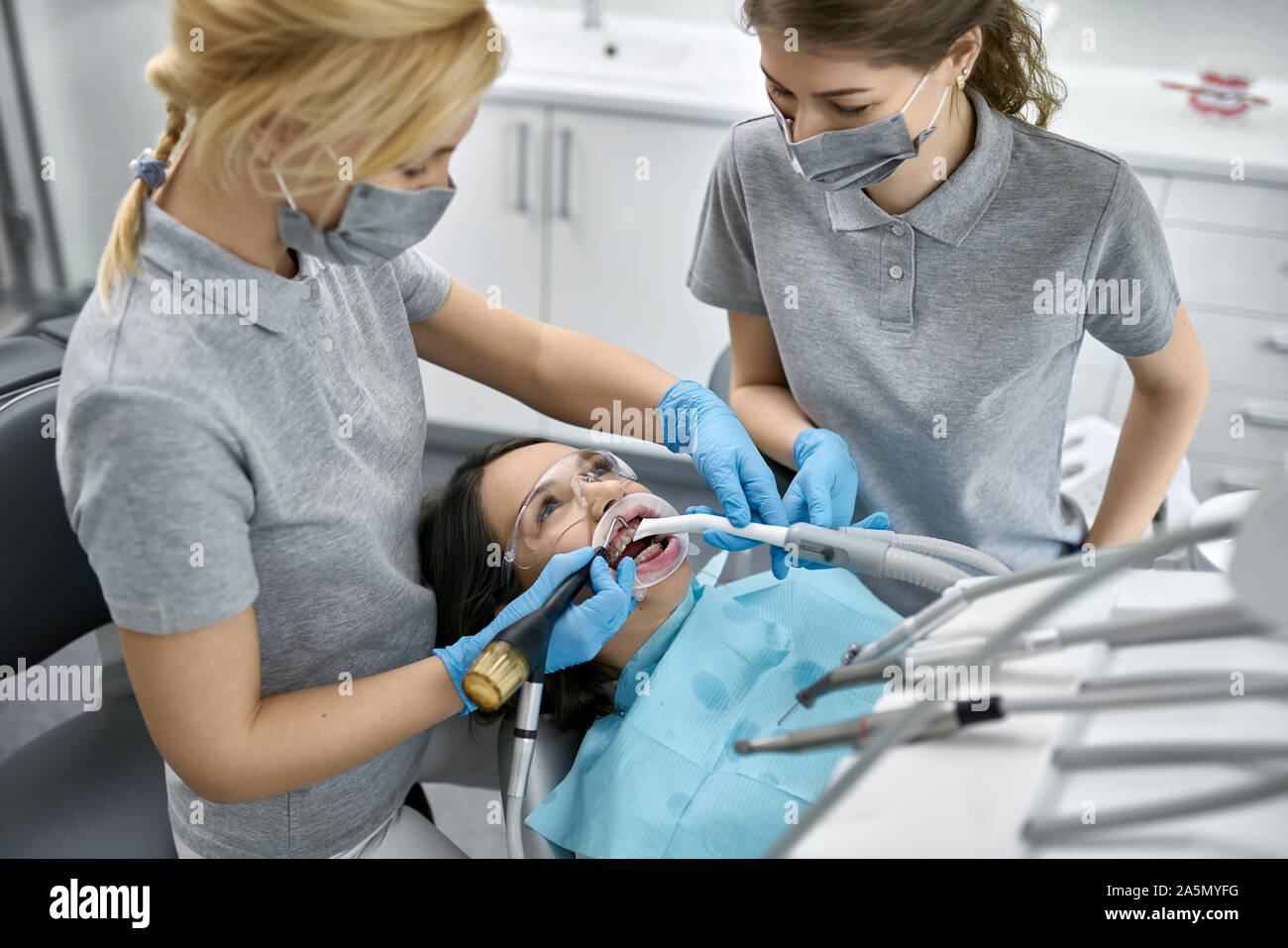 Jolie jeune fille dans des lunettes à bretelles et d'un enrouleur joue dans une clinique dentaire. Dentiste et son assistant sont nettoyer ses dents avec une il Banque D'Images Jolie jeune fille dans des lunettes à bretelles et d'un enrouleur joue dans une clinique dentaire. Dentiste et son assistant sont nettoyer ses dents avec une il Banque D'Images