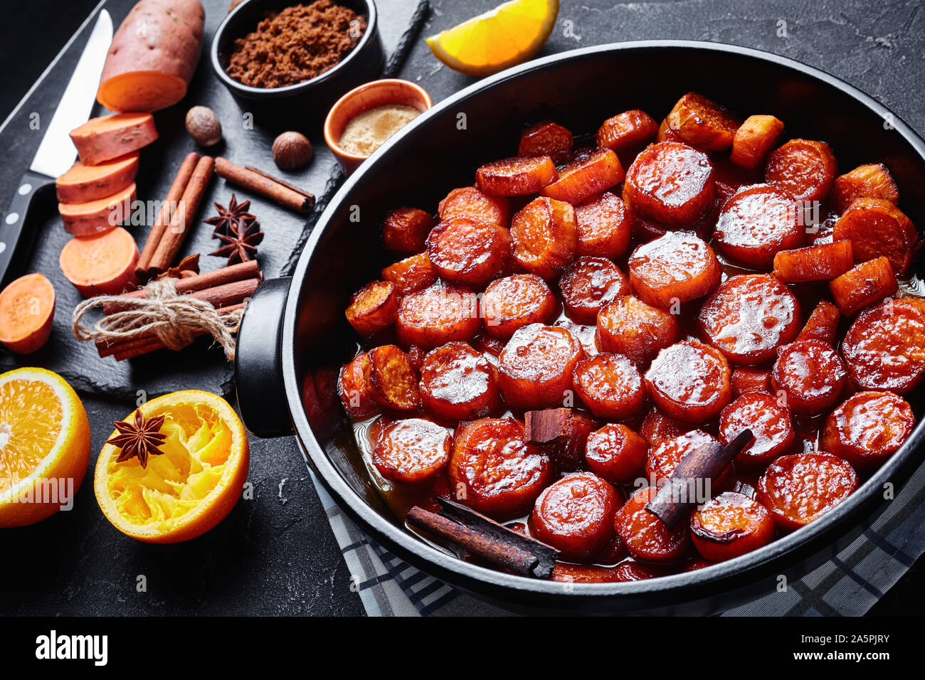 Confit d'ignames, patates douces cuites avec la cannelle, la cassonade et le beurre dans un plat de céramique noire. Ingrédients sur une liste de sélection sur une table en béton, Banque D'Images