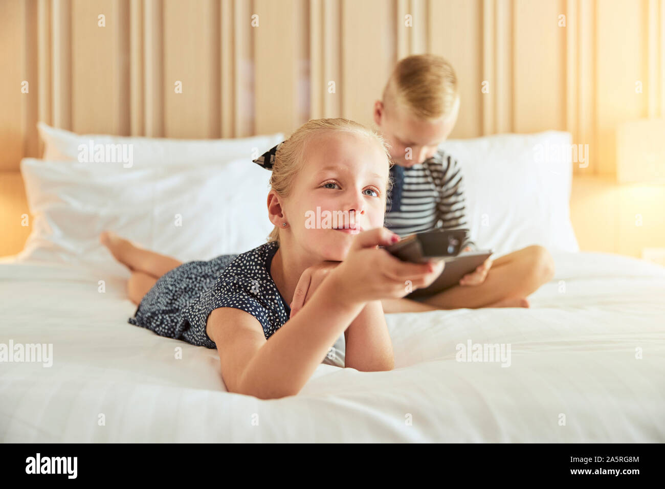 Cute little girl lying on a bed changer de chaîne pendant que vous regardez la télévision avec son frère assis derrière elle à l'aide d'une tablette numérique Banque D'Images