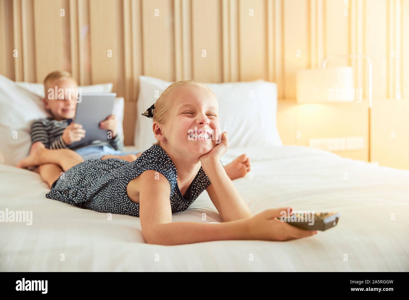 Rire petite fille allongée sur un lit à regarder la télévision avec son frère assis derrière elle à l'aide d'une tablette numérique Banque D'Images