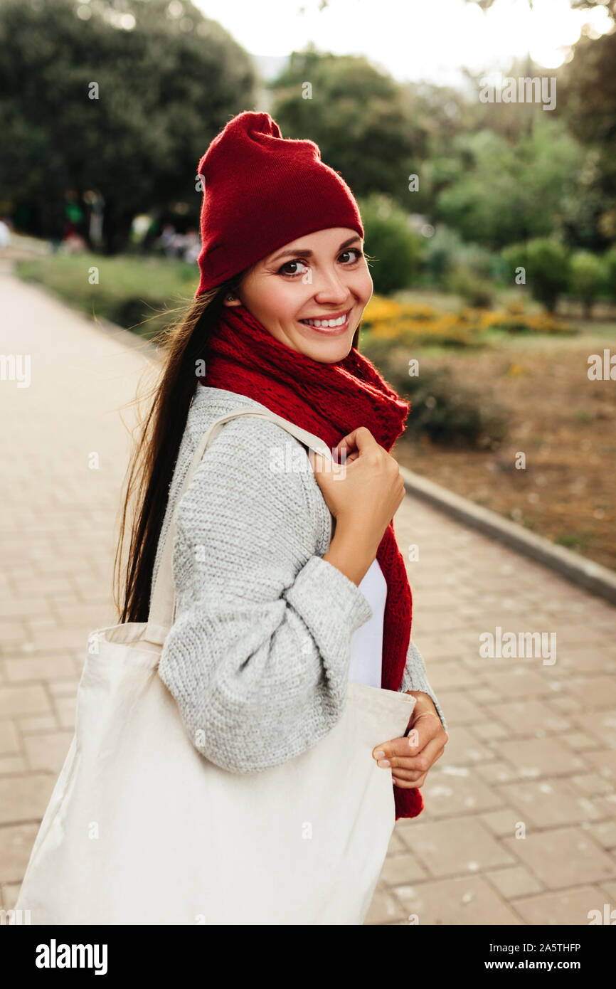 Jolie fille brune debout dans parc de la ville avec un sac sur l'épaule dans eco vêtements chauds foulard et un chapeau fashion couleurs rouges automne hiver. Banque D'Images