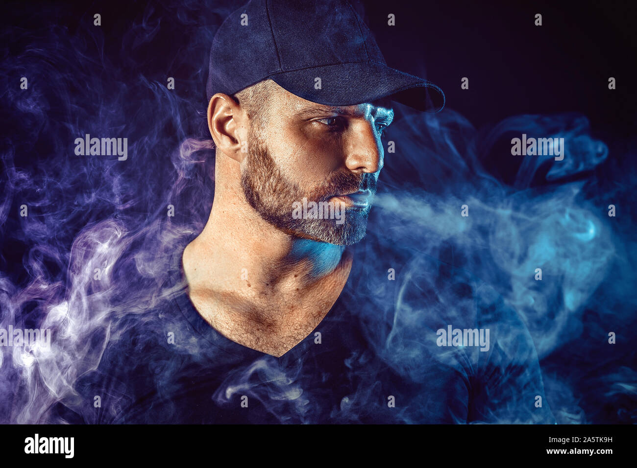 Homme barbu brutal vaping et libère un nuage de vapeur. Arrière-plan de couleur sombre. Banque D'Images