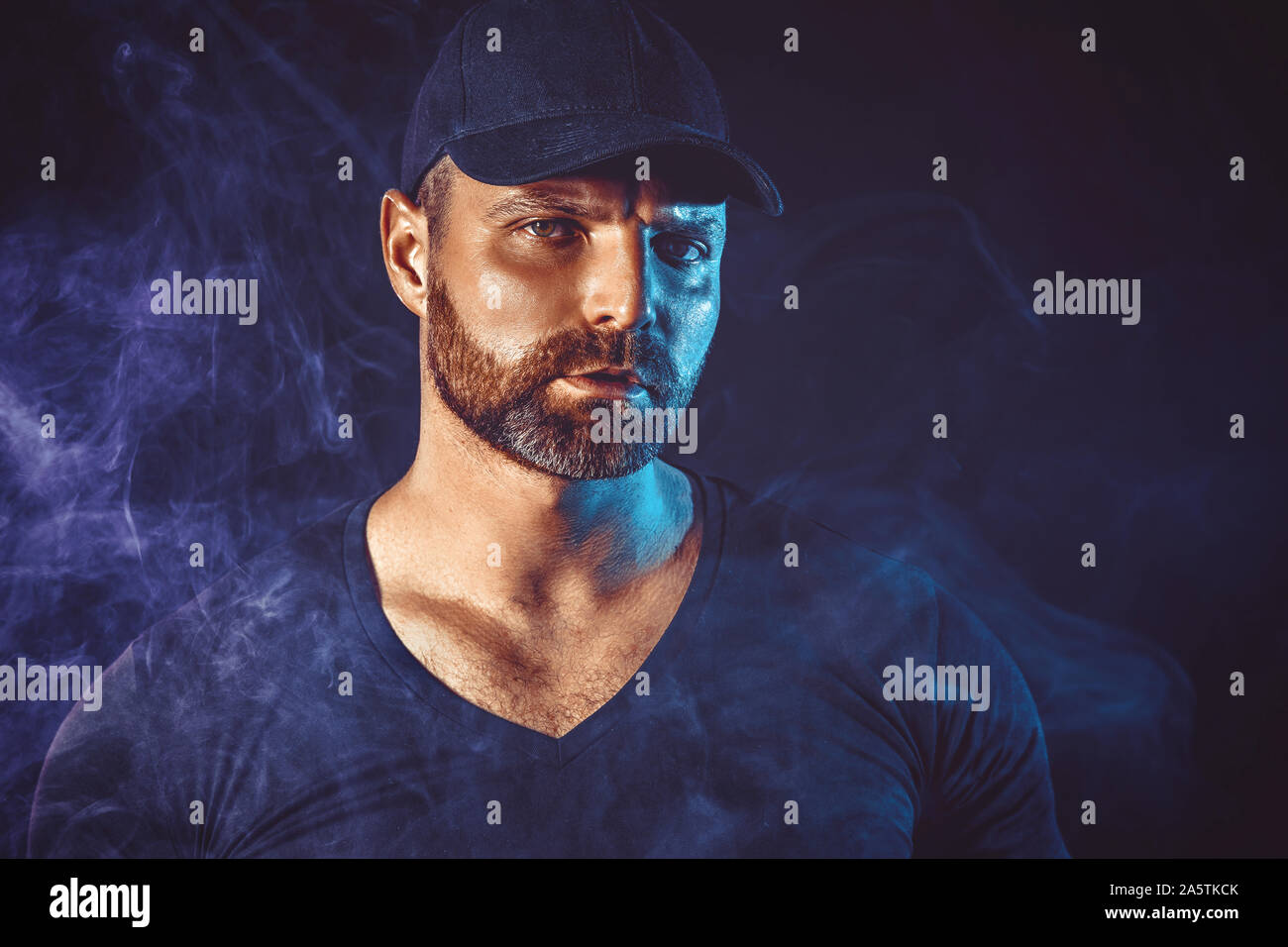 Homme barbu brutal vaping et libère un nuage de vapeur. Arrière-plan de couleur sombre. Banque D'Images