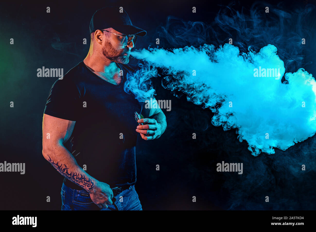Homme barbu à lunettes brutal vaping et libère un nuage de vapeur. Arrière-plan de couleur sombre avec des néons. Banque D'Images