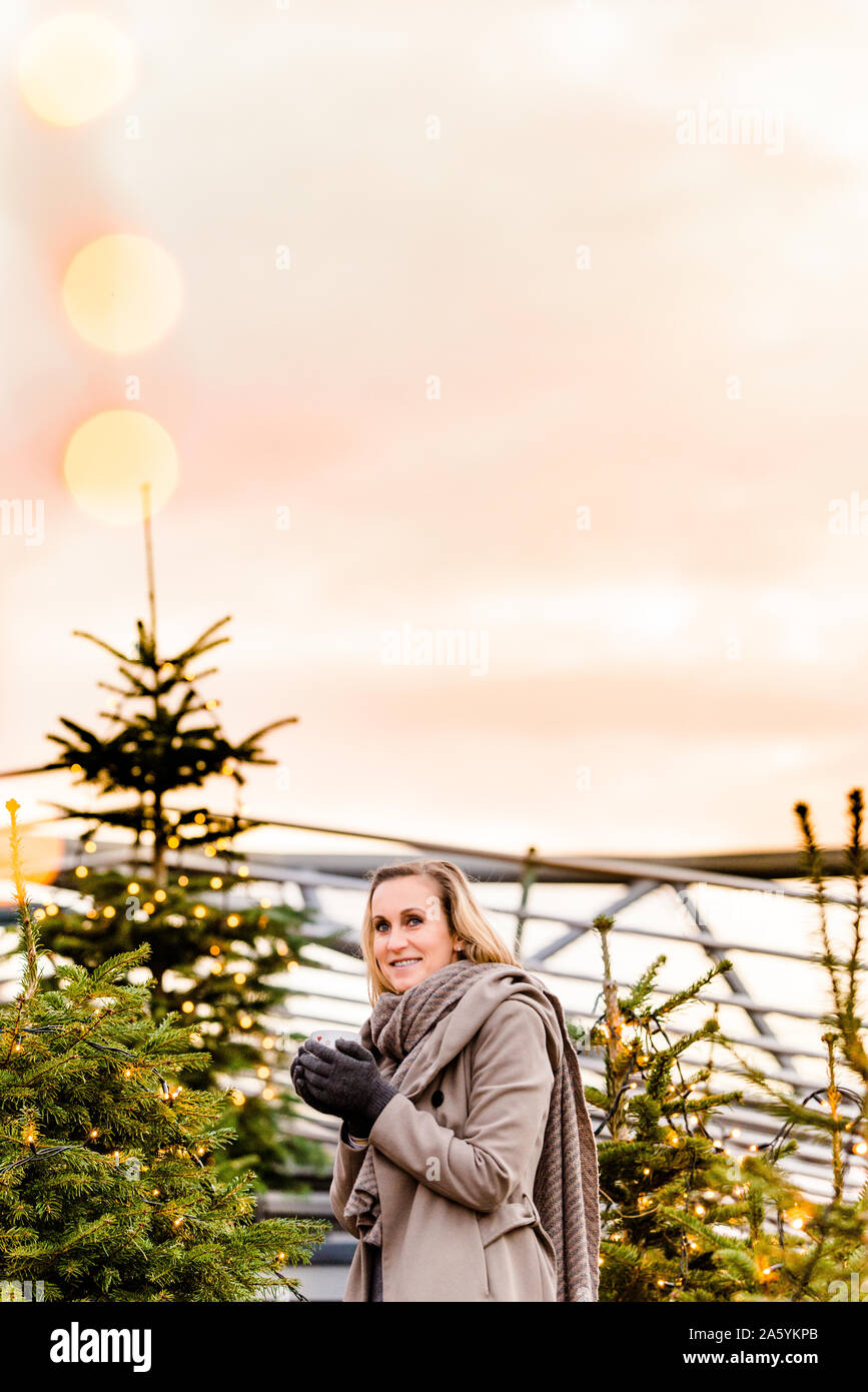 Femme tenant une boisson chaude dans ses mains à l'heure d'hiver étant entouré par des arbres de Noël Banque D'Images