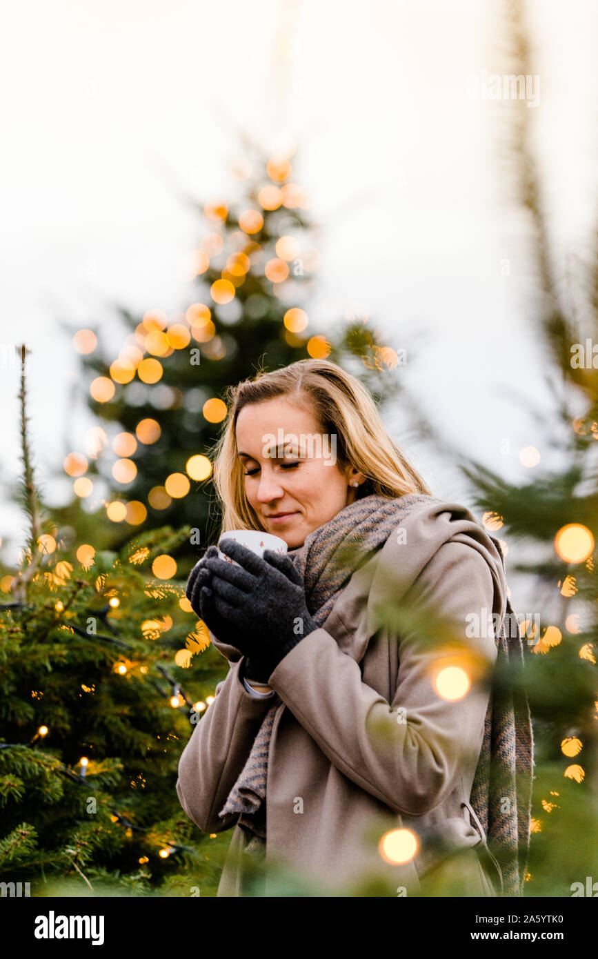 Femme tenant une boisson chaude dans ses mains à l'heure d'hiver étant entouré par des arbres de Noël Banque D'Images