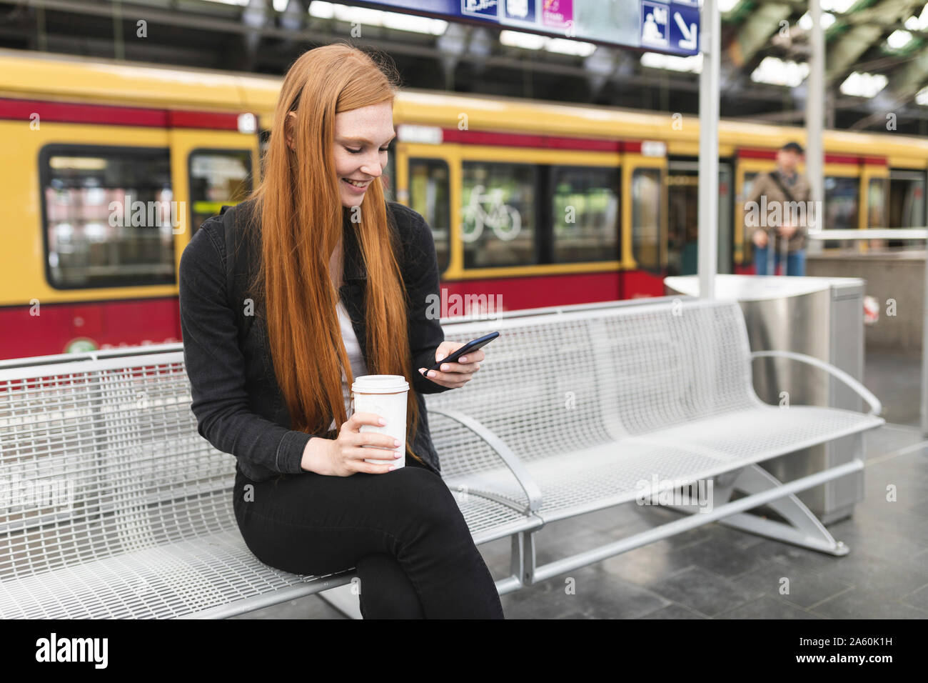 Jeune femme rousse avec du café pour aller en attente à l'aide de la plate-forme smartphone, Berlin, Allemagne Banque D'Images
