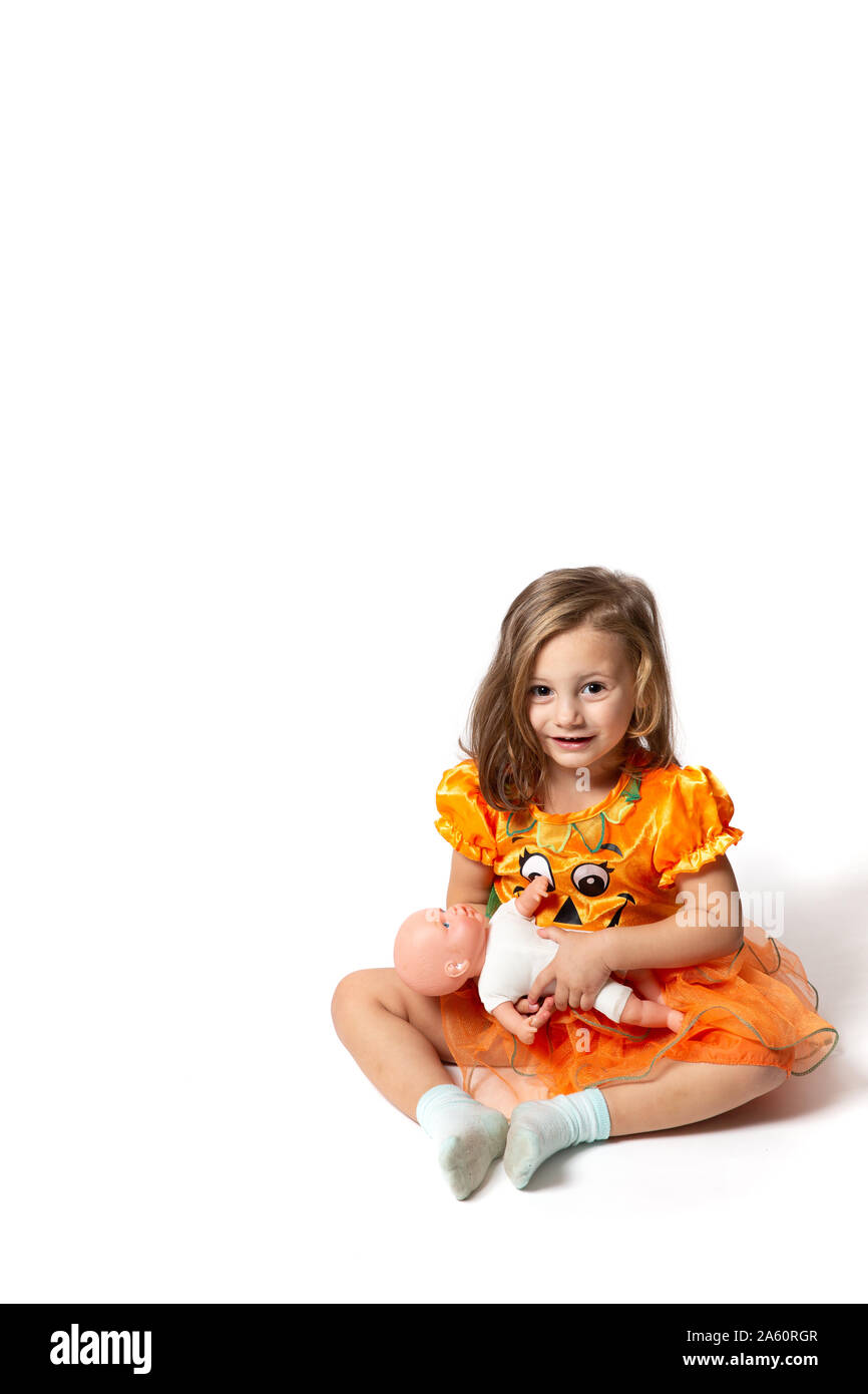 Une petite fille vêtue d'une robe orange assis sur le sol contre un fond blanc jouant avec une poupée Banque D'Images