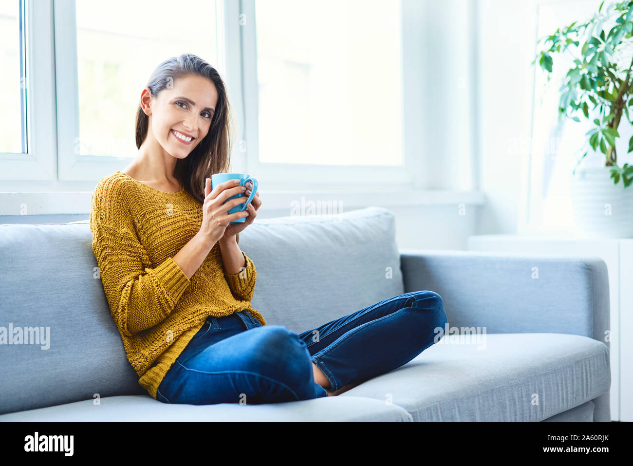 Belle jeune femme assise sur un canapé à boire du café et looking at camera Banque D'Images