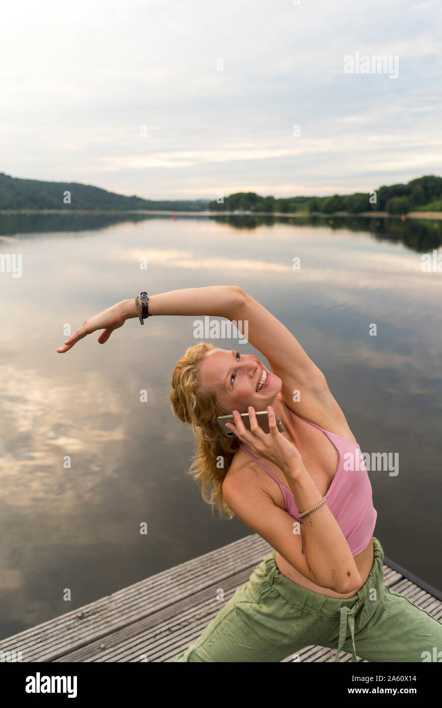 Jeune femme au téléphone faire la gymnastique sur un ponton au bord d'un lac Banque D'Images