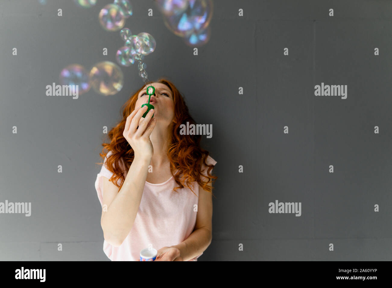 Redheaded woman leaning against wall gris soufflant des bulles de savon Banque D'Images