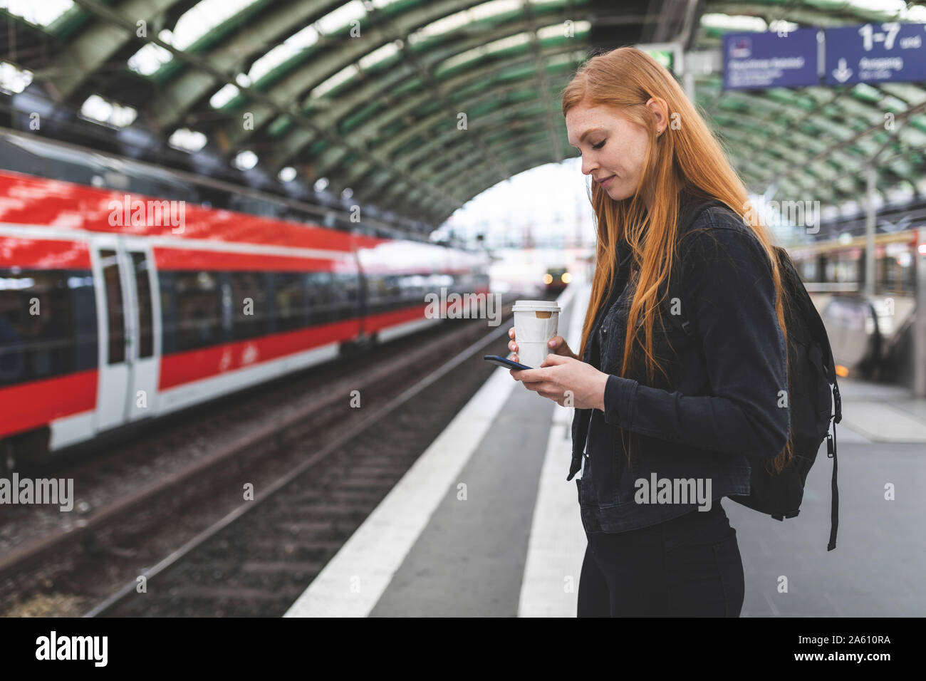 Jeune femme rousse avec du café pour aller en attente à l'aide de la plate-forme smartphone, Berlin, Allemagne Banque D'Images