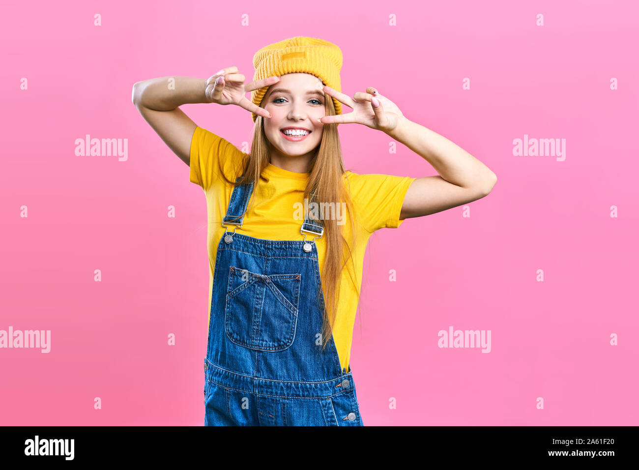 Portret une jolie fille en salopette en jean et une casquette jaune sur fond rose. Dame Fashionista student smiling . Studio lumineux de la mode la mode image Banque D'Images