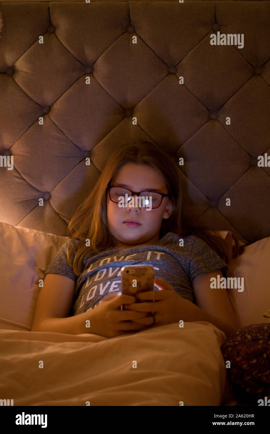 Jeune fille s'assit dans le lit en regardant son téléphone portable avant de le mettre au lit Banque D'Images