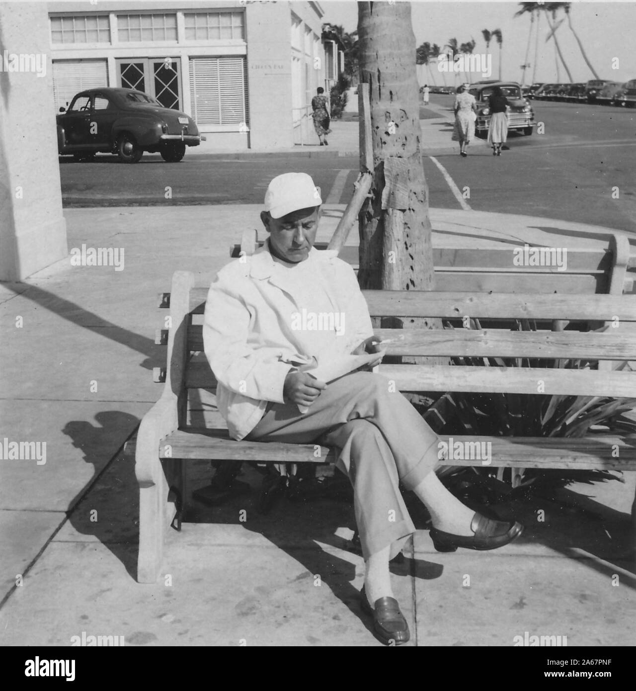 Un homme juif-américain est assis à l'extérieur sur un banc et lit un papier, avec des voitures et des magasins visibles en arrière-plan, 1940. () Banque D'Images