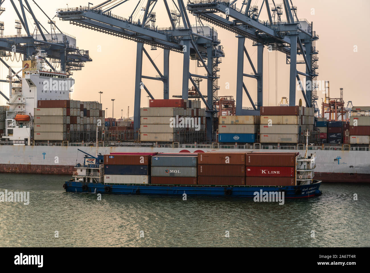 Port de Laem Chabang, Thaïlande - Mars 17, 2019 : petite Lovegistics bleu bateau passe d'apport pour conteneurs grand navire de mer sous gray grues et sunse Banque D'Images