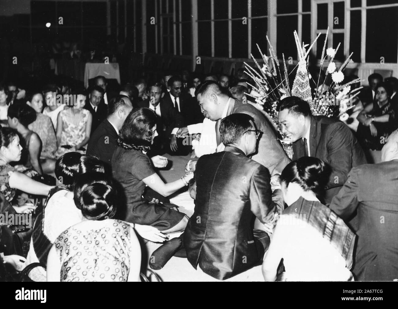 Un groupe d'hommes et de femmes habillés formellement s'assoit sur le sol, certains recevant des bracelets Sai Sin, lors d'une cérémonie Baci à une fonction conjointe de l'armée américaine et de l'armée royale thaïlandaise, photographiée pendant la guerre du Vietnam, 1968. () Banque D'Images