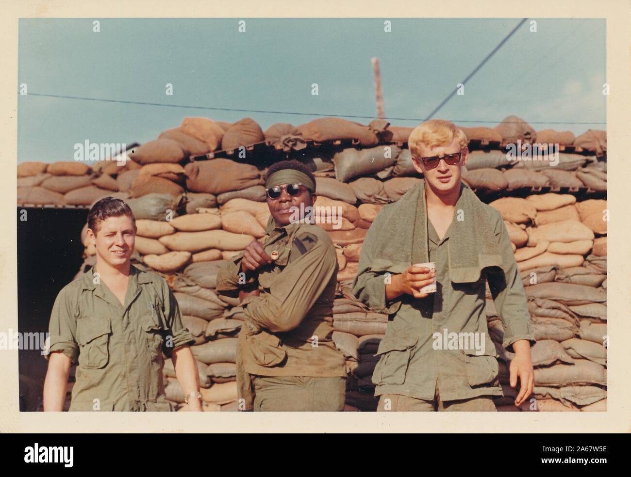 Trois jeunes militaires américains, un groupe multiracial avec deux hommes caucasiens et un homme afro-américain, sourient alors qu'ils posent devant des sacs de sable lors d'une fortification au Vietnam pendant la guerre du Vietnam, 1975. () Banque D'Images