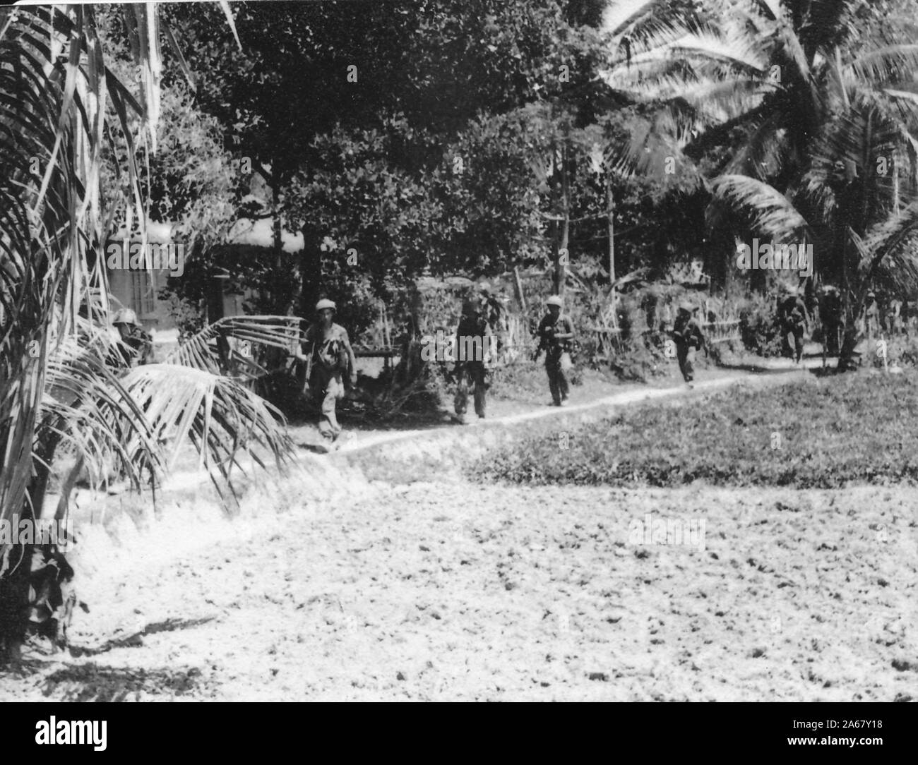 Une ligne de militaires armés, en hélices et en uniforme marchent sur un chemin de terre, un jour ensoleillé, entre un mur de village et des champs, Vietnam, 1965. () Banque D'Images