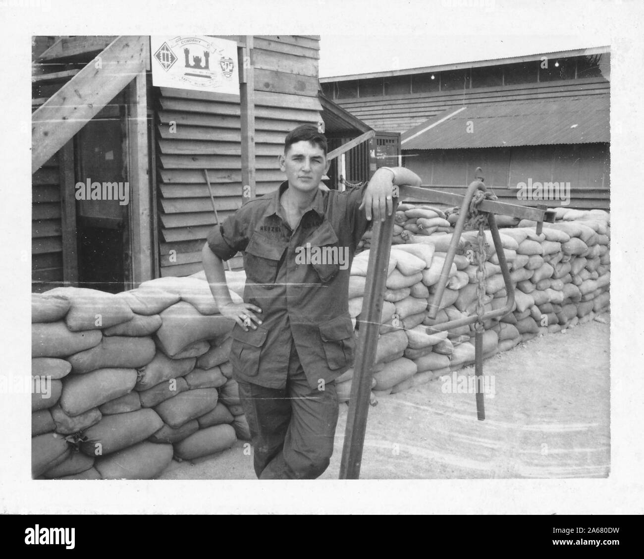 Mi-shot d'un jeune militaire américain, face à la caméra avec une expression sérieuse sur son visage, se penchant sur le bord d'un poteau tenant un triangle, près de sacs de sable et un bâtiment en bois avec un signe "Compagnie C", Vietnam, 1965. () Banque D'Images