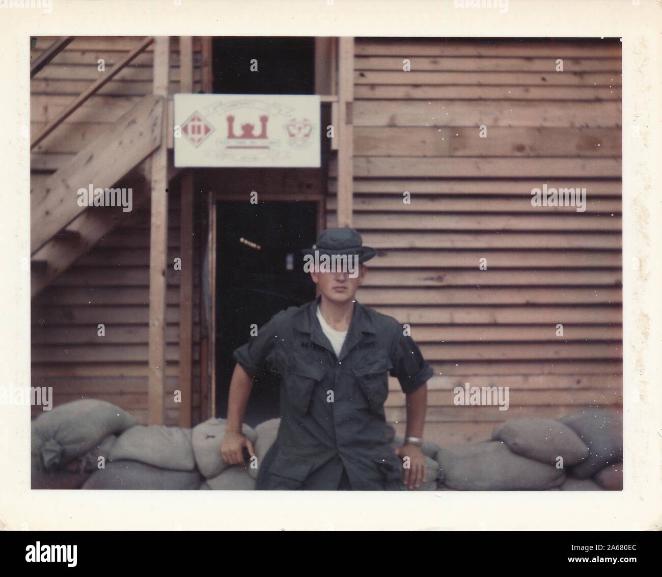 Mi-shot de Purple Heart destinataire SPC Gary Heizer, face à la caméra avec une expression sérieuse sur son visage, se penchant sur un bas mur de sacs de sable devant un bâtiment en bois avec un signe "entreprise C", Vietnam, 1965. () Banque D'Images