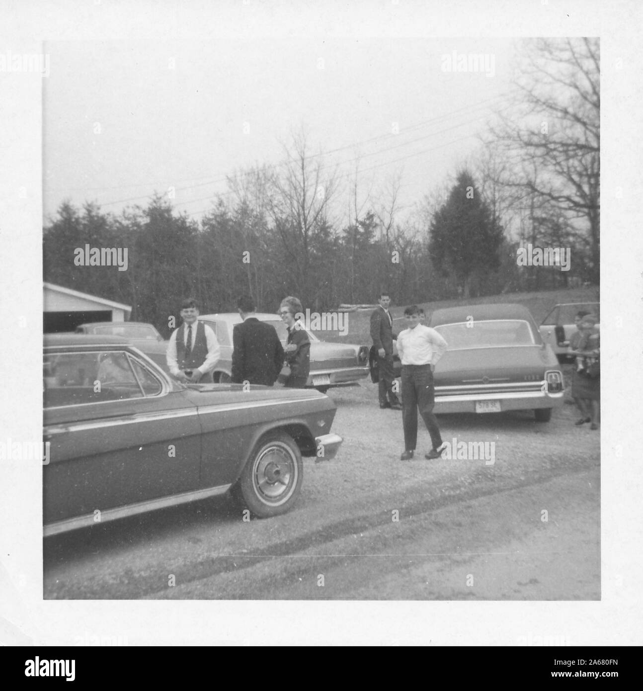 Habillez officiellement des gens, probablement des bourgers lors de funérailles militaires, tenez-vous et discutez dans une zone de banlieue herbeuse entourée de voitures anciennes, en une journée surmoulée, États-Unis d'Amérique, 1965. () Banque D'Images