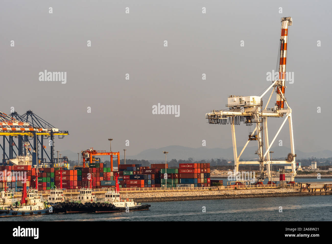 Port de Laem Chabang, Thaïlande - Mars 17, 2019 : grand récipient rouge et blanc crane au repos sur le quai rempli de piles de boîtes colorées. Peu de remorqueur Banque D'Images