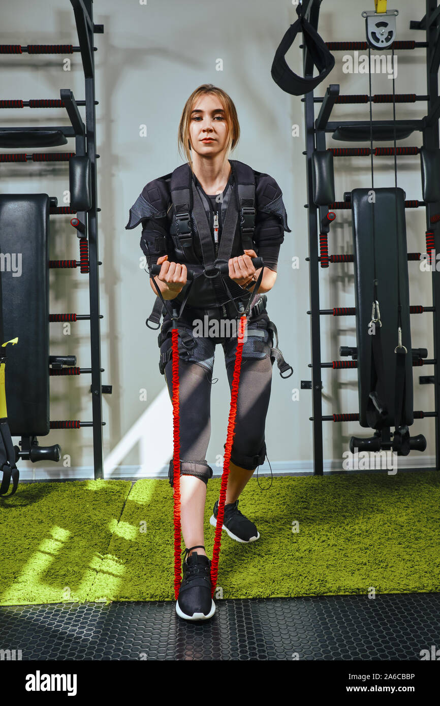 Belle dame blonde en portant des capteurs et des ceintures, se fixe avec du velcro pratique EMS fitness dans l'exécution d'exercices de gym, de traction à l'aide d'un rouge Banque D'Images